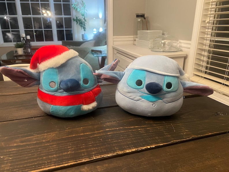 May include: Two plush toys shaped like a character with large ears. One is blue with a red Santa hat and scarf. The other is light blue with a winter hat and scarf. Both have large eyes and a dark blue nose.