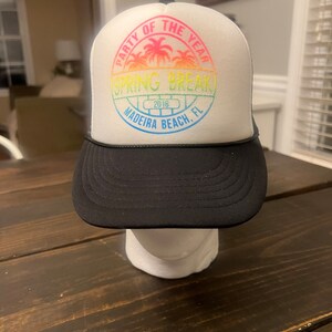 May include: White trucker hat with a black brim. The front of the hat has a colorful circular patch with the text "Party of the Year Spring Break 2016 Madeira Beach, FL".