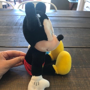 May include: A black and white plush Mickey Mouse toy sitting with yellow shoes. The toy is wearing red shorts.