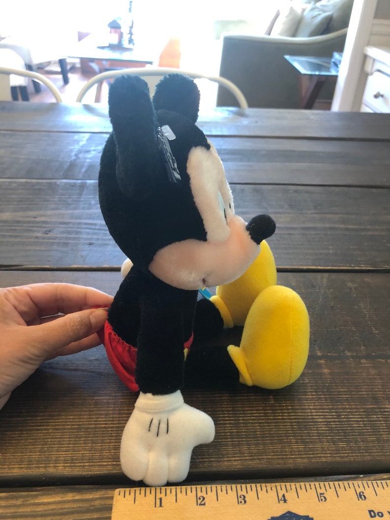 May include: A black and white plush Mickey Mouse toy sitting with yellow shoes. The toy is wearing red shorts.