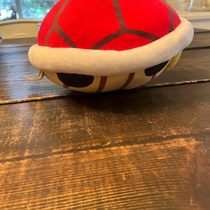 May include: A plush toy shaped like a red turtle shell with a white border. The top of the shell features a red hexagonal pattern with dark grey outlines. The underside is a light tan color with dark brown stripes and black oval markings.
