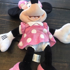 May include: Pink and white polka dot Minnie Mouse plush toy with black eyes and a big smile. The toy is wearing a pink dress with white polka dots and white shoes with pink bows. The toy is lying on a wooden surface.