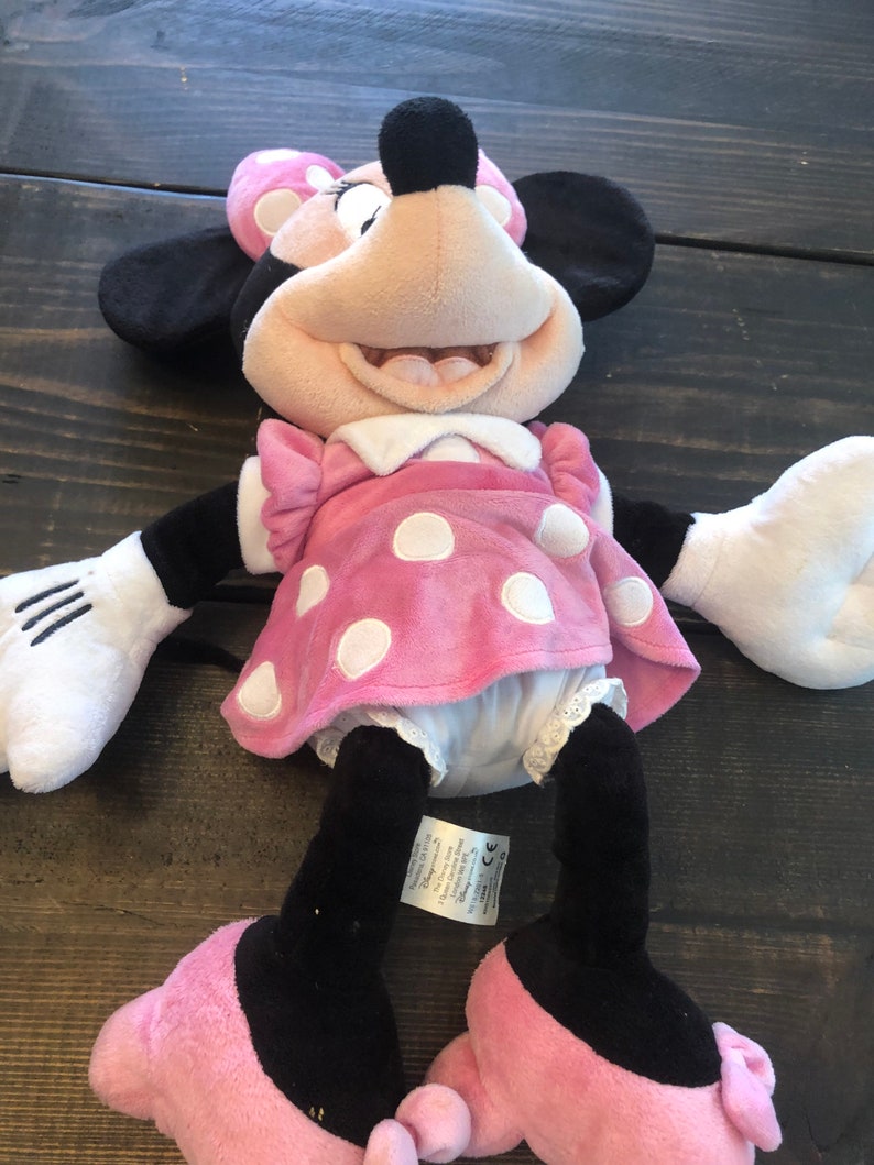 May include: Pink and white polka dot Minnie Mouse plush toy with black eyes and a big smile. The toy is wearing a pink dress with white polka dots and white shoes with pink bows. The toy is lying on a wooden surface.