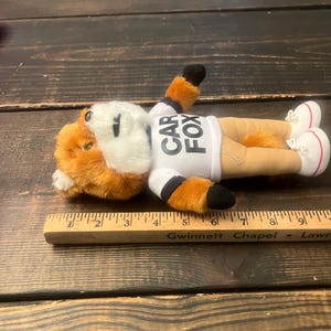 May include: A plush fox toy with orange and white fur, wearing a white shirt with "CAR FOX" printed on it. The fox is lying down, wearing tan trousers and white trainers with red accents. The toy is next to a wooden ruler.
