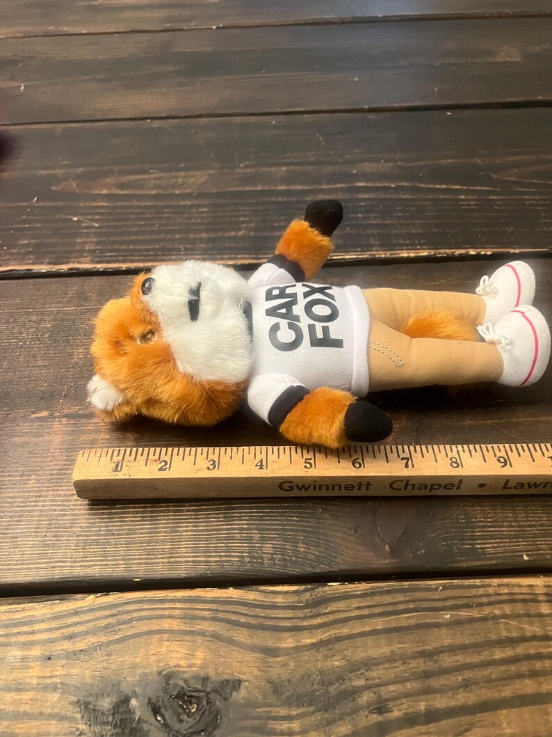 May include: A plush fox toy with orange and white fur, wearing a white shirt with "CAR FOX" printed on it. The fox is lying down, wearing tan trousers and white trainers with red accents. The toy is next to a wooden ruler.