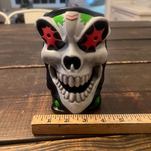 May include: A skull-shaped mug with a grinning mouth and red starburst eye designs. The mug has a black rim and green accents. A wooden ruler is placed in front of the mug. The mug is approximately 5 inches tall.