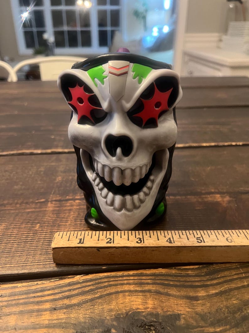 May include: A skull-shaped mug with a grinning mouth and red starburst eye designs. The mug has a black rim and green accents. A wooden ruler is placed in front of the mug. The mug is approximately 5 inches tall.