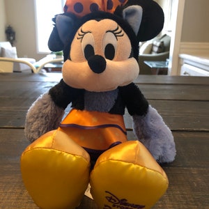 May include: A Minnie Mouse plush toy with a black head and ears, a white face, and a large orange bow with purple polka dots. The plush has an orange skirt and gold-colored feet. The Disney Store 2019 logo is on the foot.
