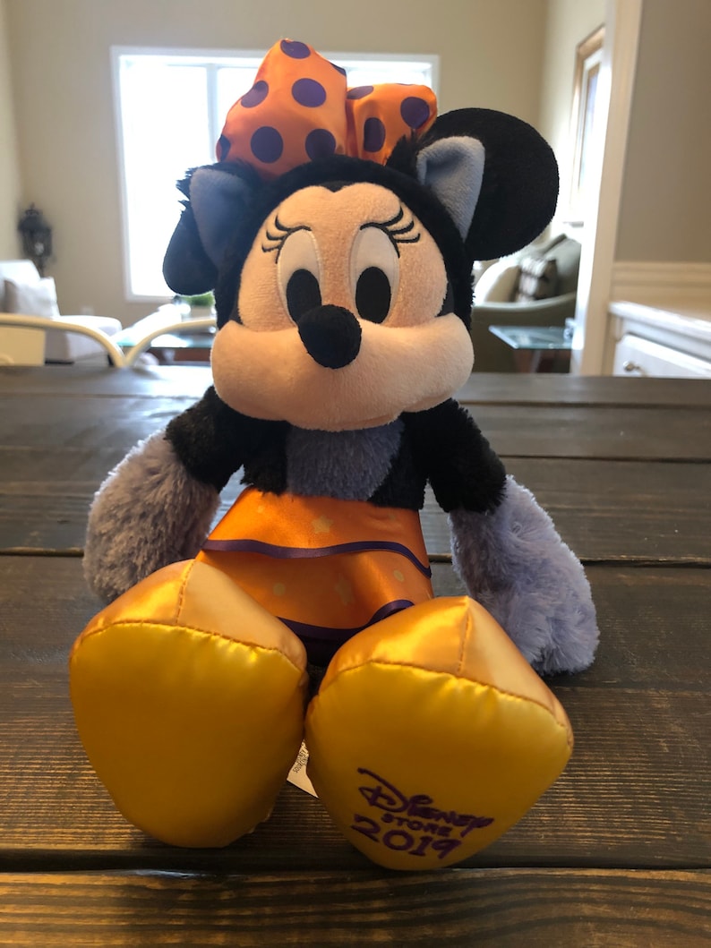 May include: A Minnie Mouse plush toy with a black head and ears, a white face, and a large orange bow with purple polka dots. The plush has an orange skirt and gold-colored feet. The Disney Store 2019 logo is on the foot.