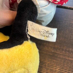 May include: A yellow and black plush toy wearing a red and white polka dot dress with white lace trim. The toy has a tag that reads "Walt Disney World."