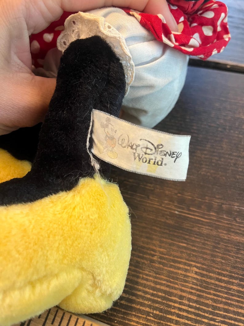 May include: A yellow and black plush toy wearing a red and white polka dot dress with white lace trim. The toy has a tag that reads "Walt Disney World."