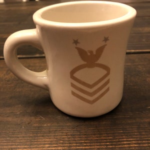 May include: White ceramic coffee mug with a gold eagle and chevron design. The mug has two stars above the eagle.