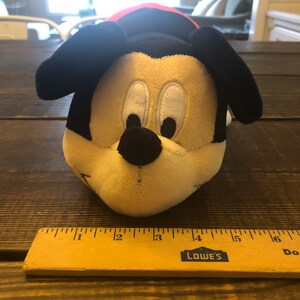 May include: A plush Mickey Mouse toy with black ears, a black nose, and white eyes. The toy is lying on a wooden surface with a ruler next to it.