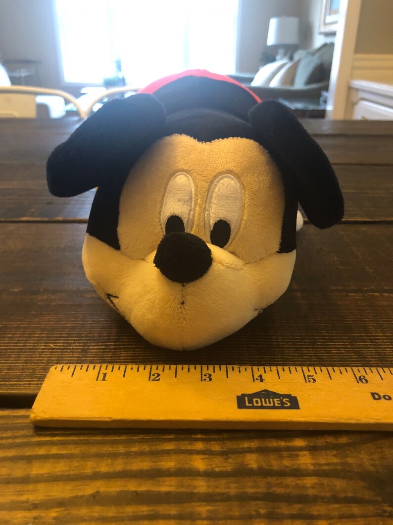 May include: A plush Mickey Mouse toy with black ears, a black nose, and white eyes. The toy is lying on a wooden surface with a ruler next to it.