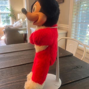 May include: A vintage Mickey Mouse plush toy, featuring a red and white outfit with black fur and a yellow face. The plush is standing on a white metal stand, showcasing the classic character's iconic design. The toy is positioned on a dark wooden table.