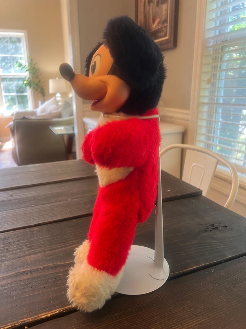 May include: A vintage Mickey Mouse plush toy, featuring a red and white outfit with black fur and a yellow face. The plush is standing on a white metal stand, showcasing the classic character's iconic design. The toy is positioned on a dark wooden table.