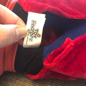 May include: Close-up of a red, blue, and black plush toy with a white tag that reads "Toy Factory" with a gear logo and the text "San Antonio, TX".