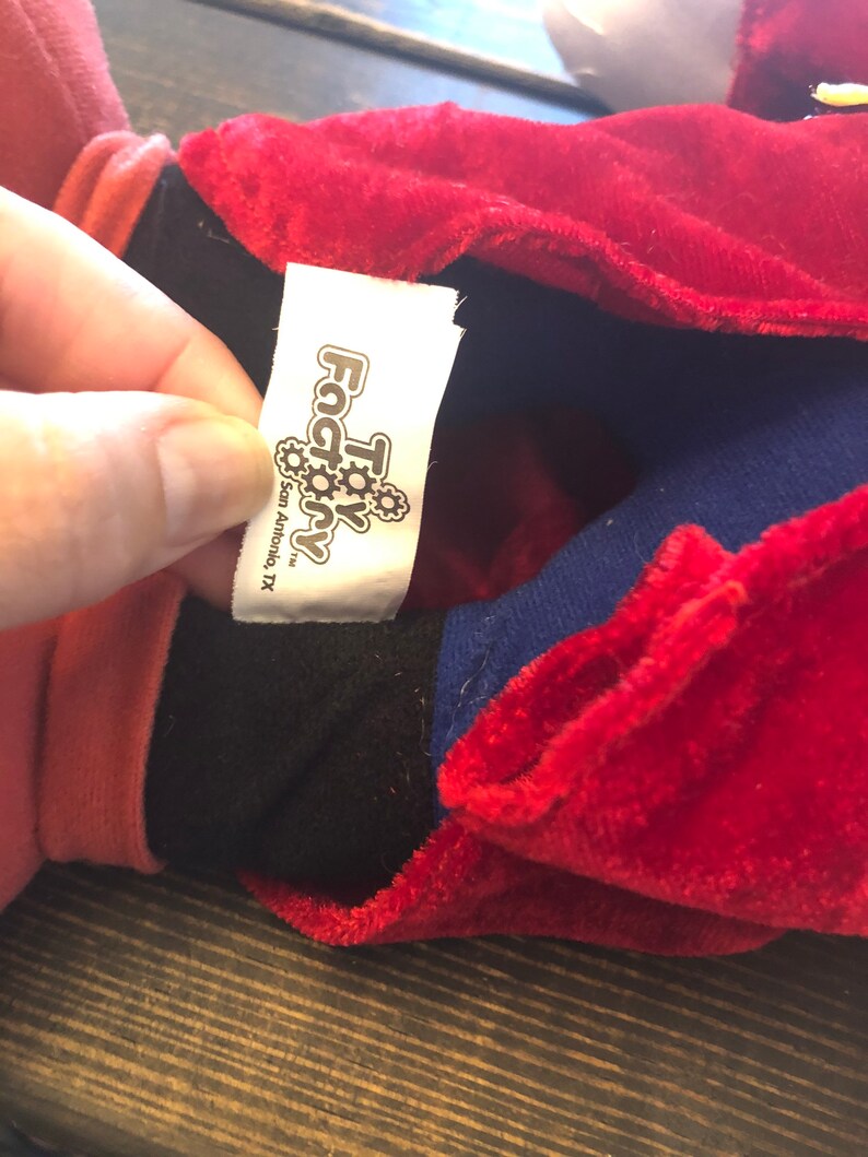 May include: Close-up of a red, blue, and black plush toy with a white tag that reads "Toy Factory" with a gear logo and the text "San Antonio, TX".