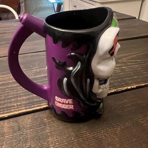 May include: Purple ceramic mug with a skull design on one side and the words "Grave Digger" in white. The mug has a black interior and a purple handle.