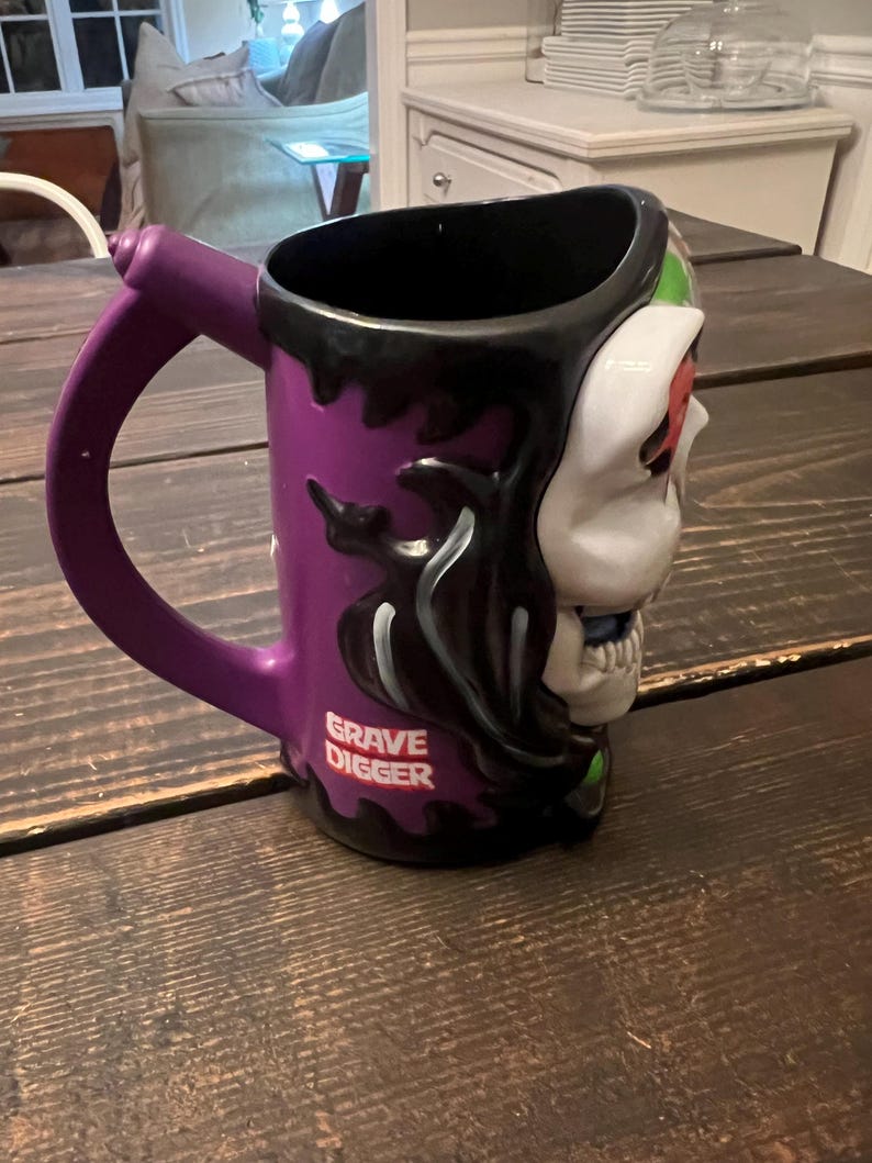 May include: Purple ceramic mug with a skull design on one side and the words "Grave Digger" in white. The mug has a black interior and a purple handle.