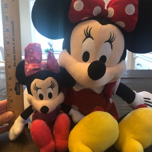 May include: Two plush Minnie Mouse toys. The larger Minnie Mouse is wearing a red dress with white polka dots and a red bow with white polka dots. The smaller Minnie Mouse is wearing a pink dress with white hearts and a pink bow with white hearts. Both Minnie Mouse toys have black ears and yellow shoes.