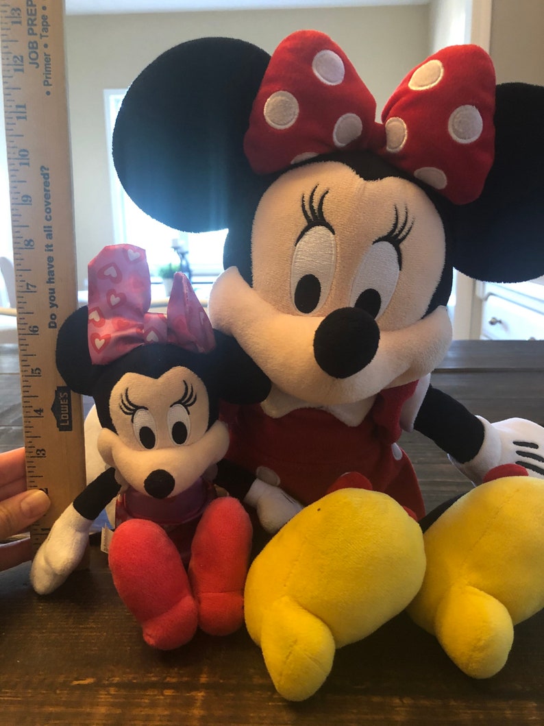May include: Two plush Minnie Mouse toys. The larger Minnie Mouse is wearing a red dress with white polka dots and a red bow with white polka dots. The smaller Minnie Mouse is wearing a pink dress with white hearts and a pink bow with white hearts. Both Minnie Mouse toys have black ears and yellow shoes.
