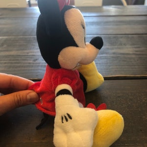 Op de afbeelding: Een pluche speelgoed van Minnie Mouse die een rode jurk draagt met witte stippen en een rode strik. Ze heeft zwarte oren, een zwarte neus en witte handschoenen. Haar voeten zijn geel.