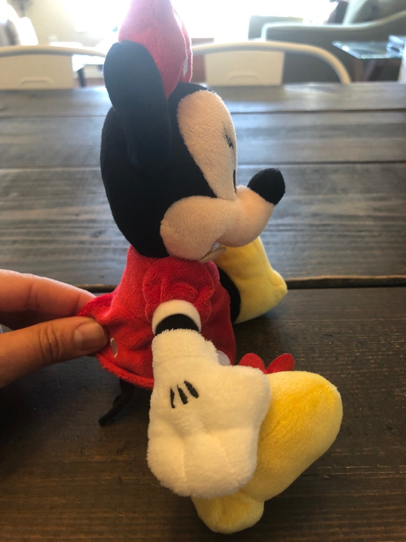Op de afbeelding: Een pluche speelgoed van Minnie Mouse die een rode jurk draagt met witte stippen en een rode strik. Ze heeft zwarte oren, een zwarte neus en witte handschoenen. Haar voeten zijn geel.