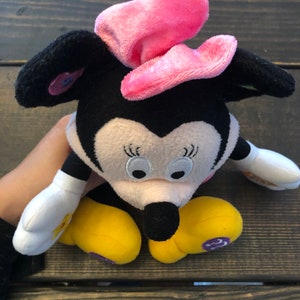 May include: A plush black and white Minnie Mouse toy with a pink bow. The toy has yellow shoes and a white shirt with a flower design.