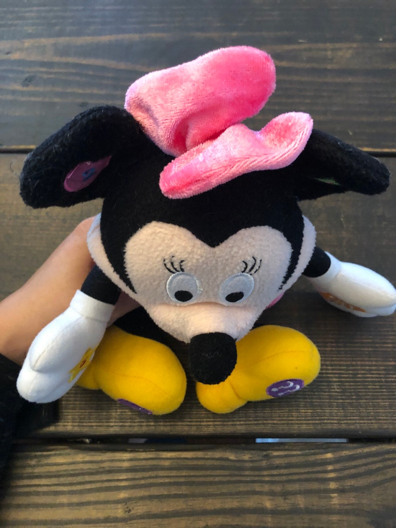May include: A plush black and white Minnie Mouse toy with a pink bow. The toy has yellow shoes and a white shirt with a flower design.