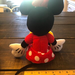 May include: A plush toy of Minnie Mouse wearing a red dress with white polka dots and a yellow bow. The toy is sitting on a wooden surface with a ruler in the foreground.