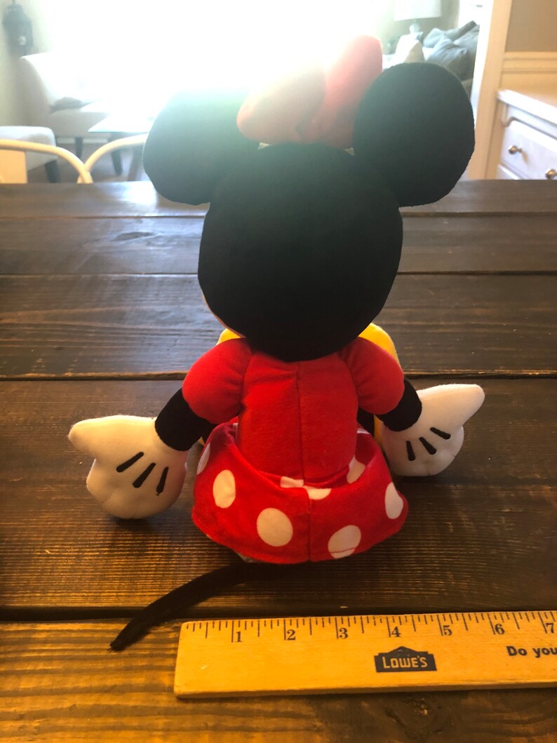 May include: A plush toy of Minnie Mouse wearing a red dress with white polka dots and a yellow bow. The toy is sitting on a wooden surface with a ruler in the foreground.