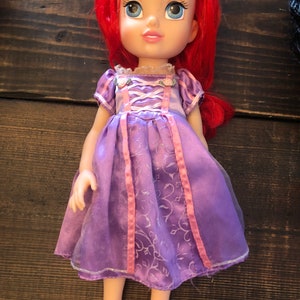 May include: A purple and pink dress with a floral pattern and pink ribbon accents. The doll has long red hair and blue eyes.
