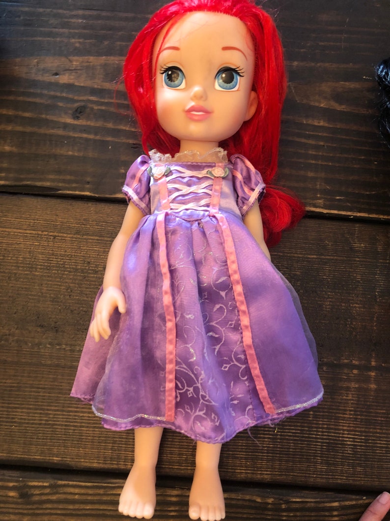 May include: A purple and pink dress with a floral pattern and pink ribbon accents. The doll has long red hair and blue eyes.