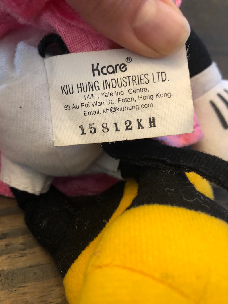 May include: A white, pink, and yellow plush toy with a black tag that reads "Kcare KIU HUNG INDUSTRIES LTD. 14/F., Yale Ind. Centre, 63 Au Pui Wan St., Fotan, Hong Kong. Email: kh@kiuhung.com 15812KH"