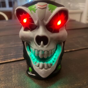 May include: A skull-shaped decorative item with glowing red eyes and a green illuminated mouth. The skull is white with black and green accents. The item is sitting on a dark wooden surface.