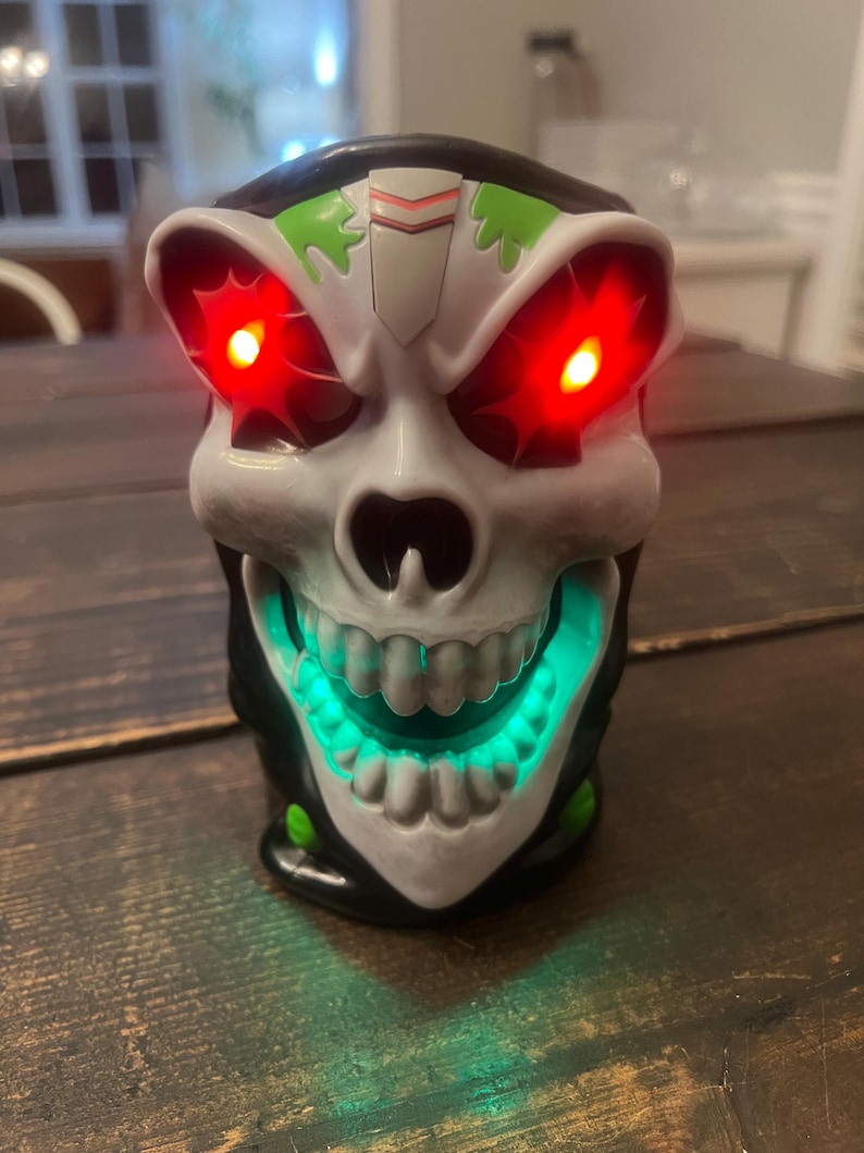 May include: A skull-shaped decorative item with glowing red eyes and a green illuminated mouth. The skull is white with black and green accents. The item is sitting on a dark wooden surface.