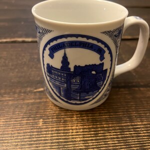 May include: White ceramic mug with a blue and white design featuring a cityscape and the text "PHILADELPHIA, PA".