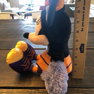 May include: A plush Minnie Mouse doll in a seated position, featuring a black head and body, a yellow face, and a purple and orange outfit. The doll has a bow with orange and purple polka dots. A ruler shows the doll is approximately 9 inches tall.