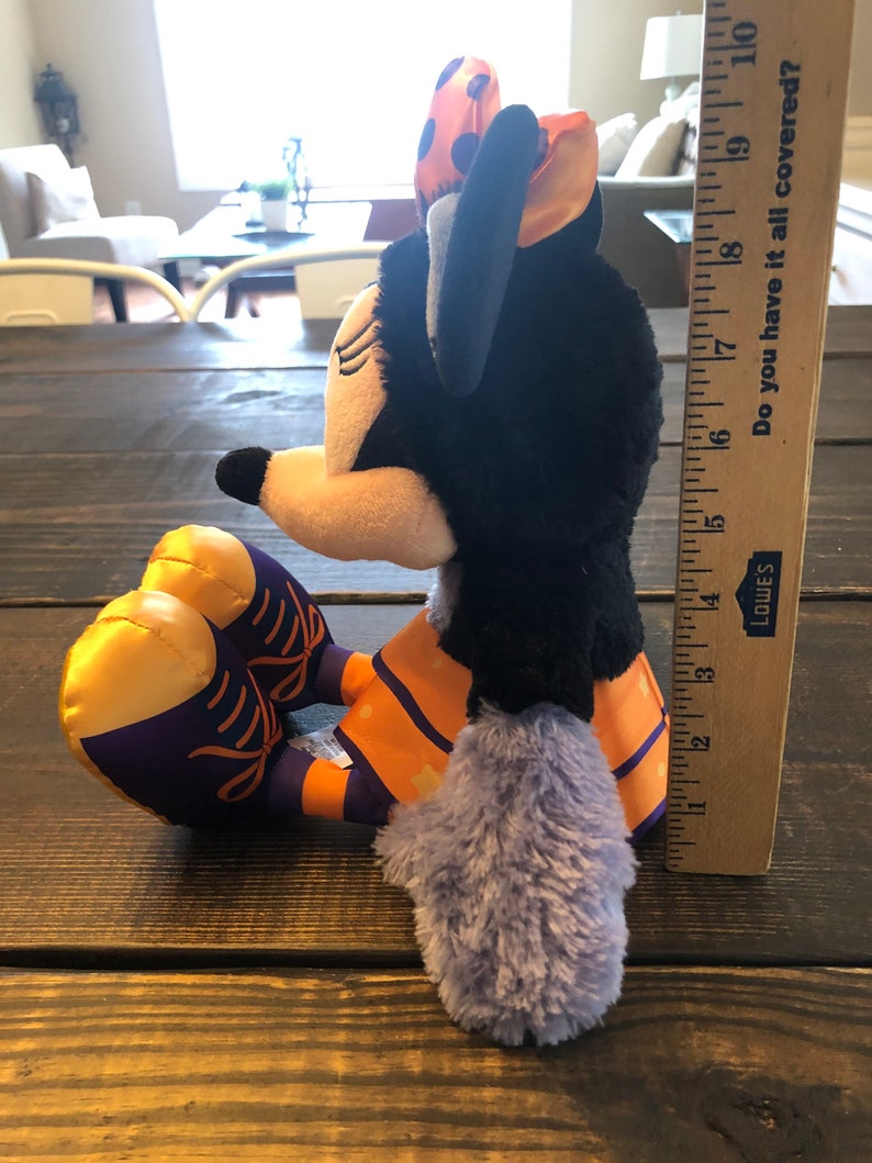 May include: A plush Minnie Mouse doll in a seated position, featuring a black head and body, a yellow face, and a purple and orange outfit. The doll has a bow with orange and purple polka dots. A ruler shows the doll is approximately 9 inches tall.