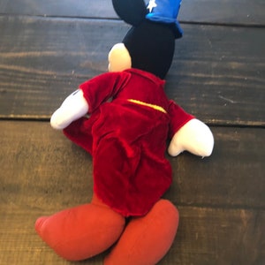 May include: A plush Mickey Mouse toy wearing a red robe and a blue wizard hat with white stars. The toy is standing on a wooden surface.