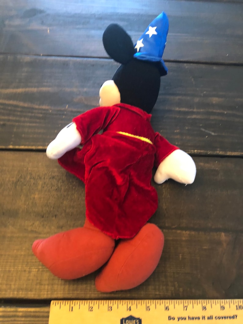 May include: A plush Mickey Mouse toy wearing a red robe and a blue wizard hat with white stars. The toy is standing on a wooden surface.