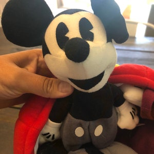 Peut inclure: Peluche Mickey Mouse noir et blanc avec un tissu rouge et violet en arri&egrave;re-plan. Le jouet a un nez noir, des yeux blancs et un grand sourire.