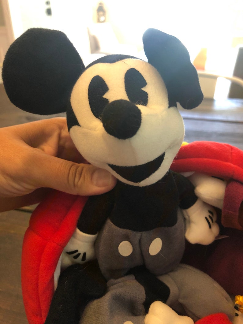 Peut inclure: Peluche Mickey Mouse noir et blanc avec un tissu rouge et violet en arri&egrave;re-plan. Le jouet a un nez noir, des yeux blancs et un grand sourire.