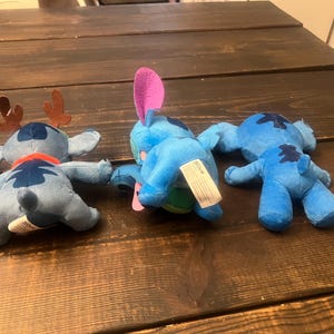 Op de afbeelding: Drie blauwe pluche speelgoed van het personage Stitch uit Lilo & Stitch, liggend op een donker houten oppervlak. E&eacute;n heeft bruine geweien en een rode sjaal, een ander een roze oor, en de derde is effen.