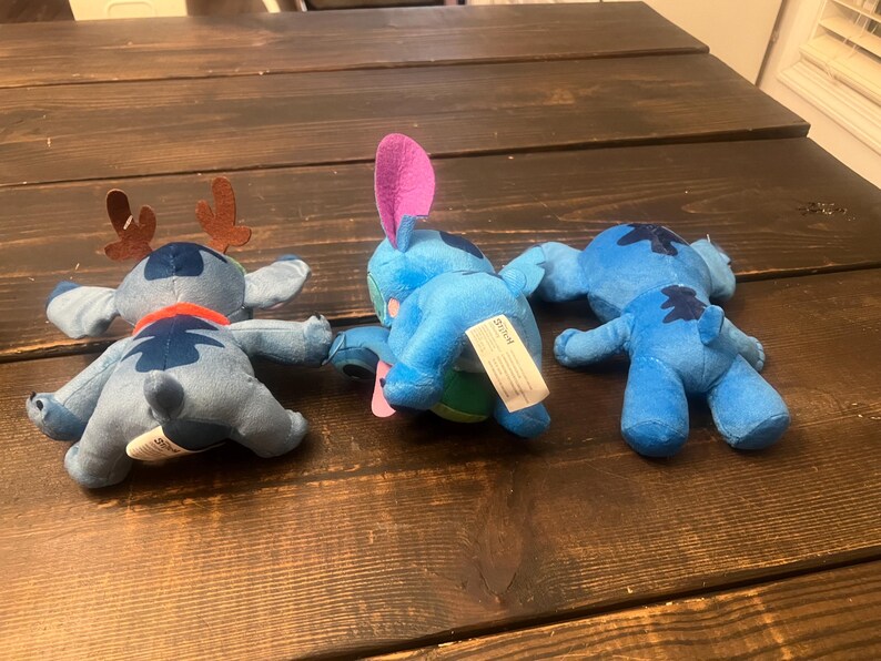 Op de afbeelding: Drie blauwe pluche speelgoed van het personage Stitch uit Lilo & Stitch, liggend op een donker houten oppervlak. E&eacute;n heeft bruine geweien en een rode sjaal, een ander een roze oor, en de derde is effen.