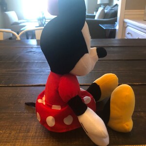 May include: A red and white polka dot dress with a black bow on top of the head. The plush toy is sitting on a wooden surface with a ruler in the foreground. The ruler is marked with inches and has the word "LOWE'S" printed on it.