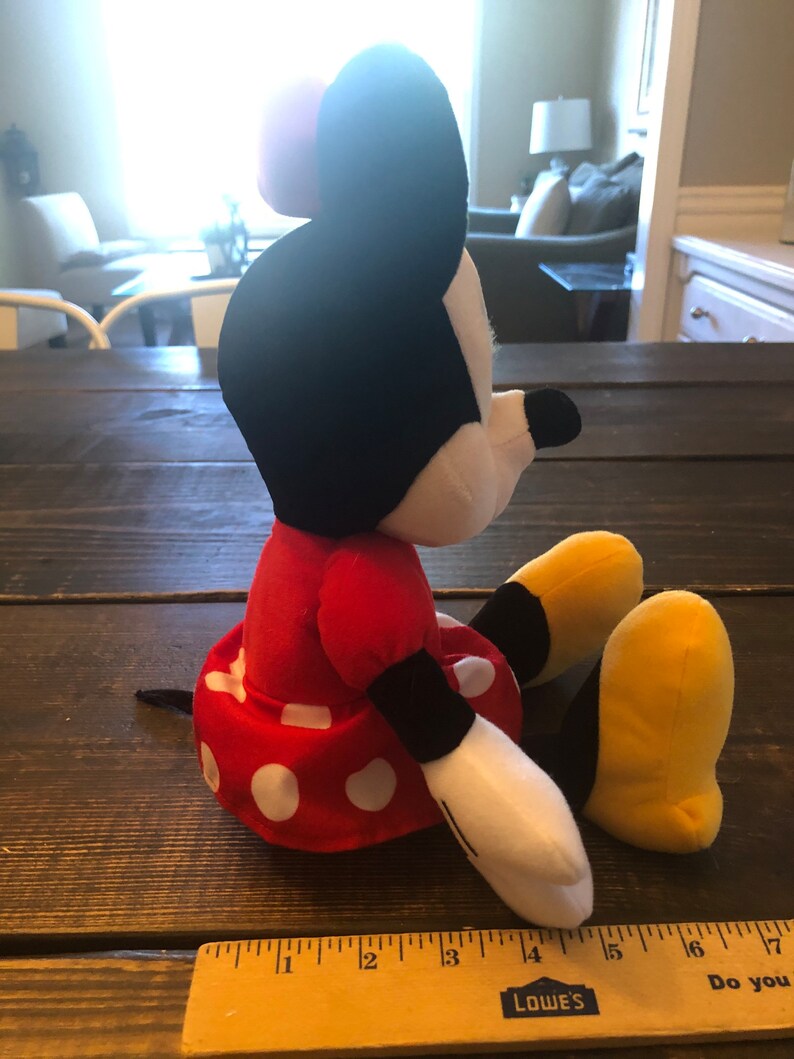 May include: A red and white polka dot dress with a black bow on top of the head. The plush toy is sitting on a wooden surface with a ruler in the foreground. The ruler is marked with inches and has the word "LOWE'S" printed on it.