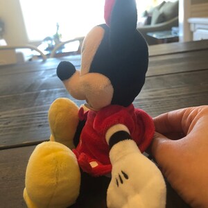 Op de afbeelding: Een pluche speelgoed van Minnie Mouse, zittend met haar benen gekruist. Ze draagt een rode jurk met witte stippen en gele schoenen. Haar oren zijn rood en zwart, en haar gezicht is wit met zwarte ogen en een zwarte neus.