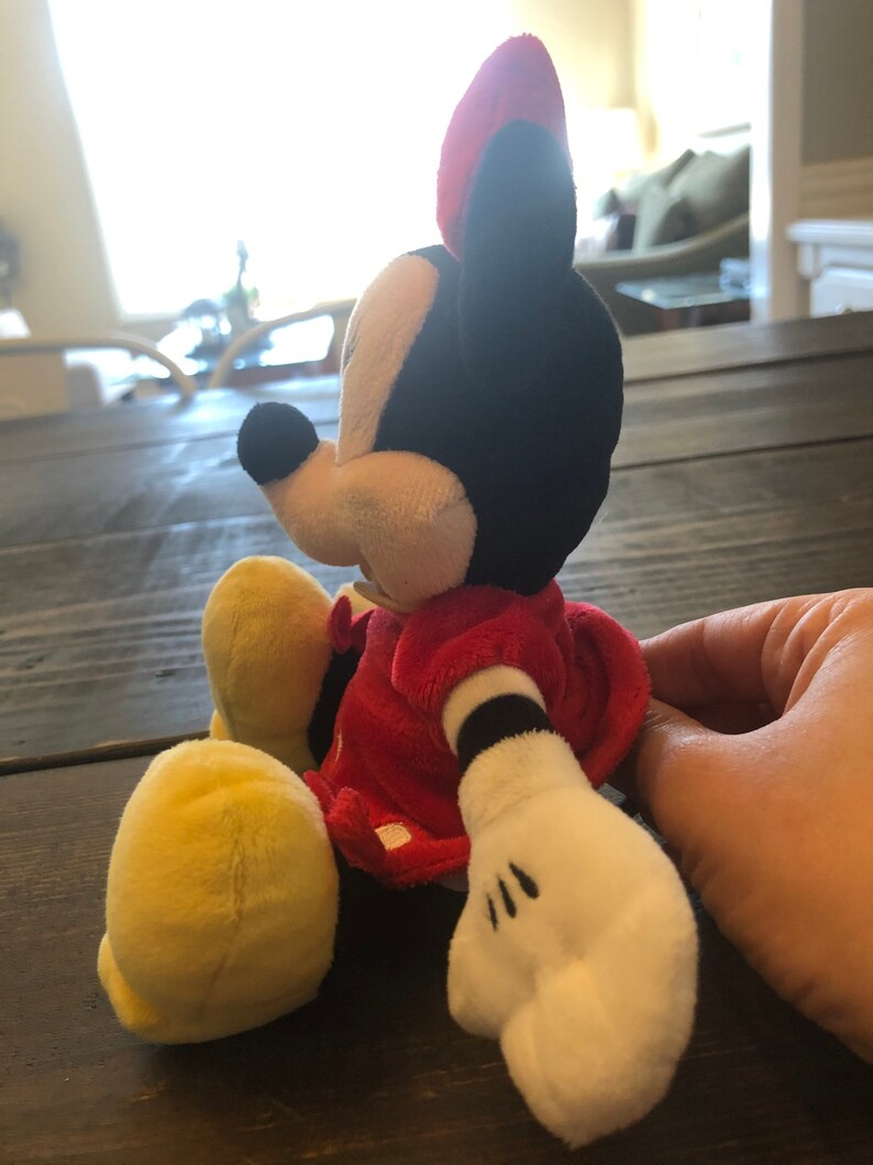 Op de afbeelding: Een pluche speelgoed van Minnie Mouse, zittend met haar benen gekruist. Ze draagt een rode jurk met witte stippen en gele schoenen. Haar oren zijn rood en zwart, en haar gezicht is wit met zwarte ogen en een zwarte neus.